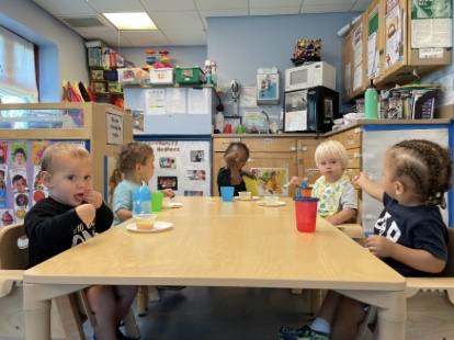 Six toddler aged children sitting around a big rectangular table in a classroom.